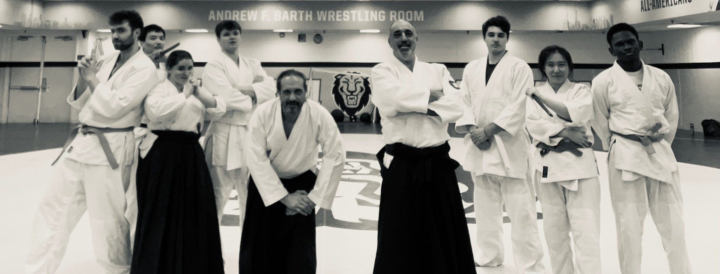 Nine club members in white gi, three in black hakama, standing together on the mat in front of the Columbia lion in the university wrestling room